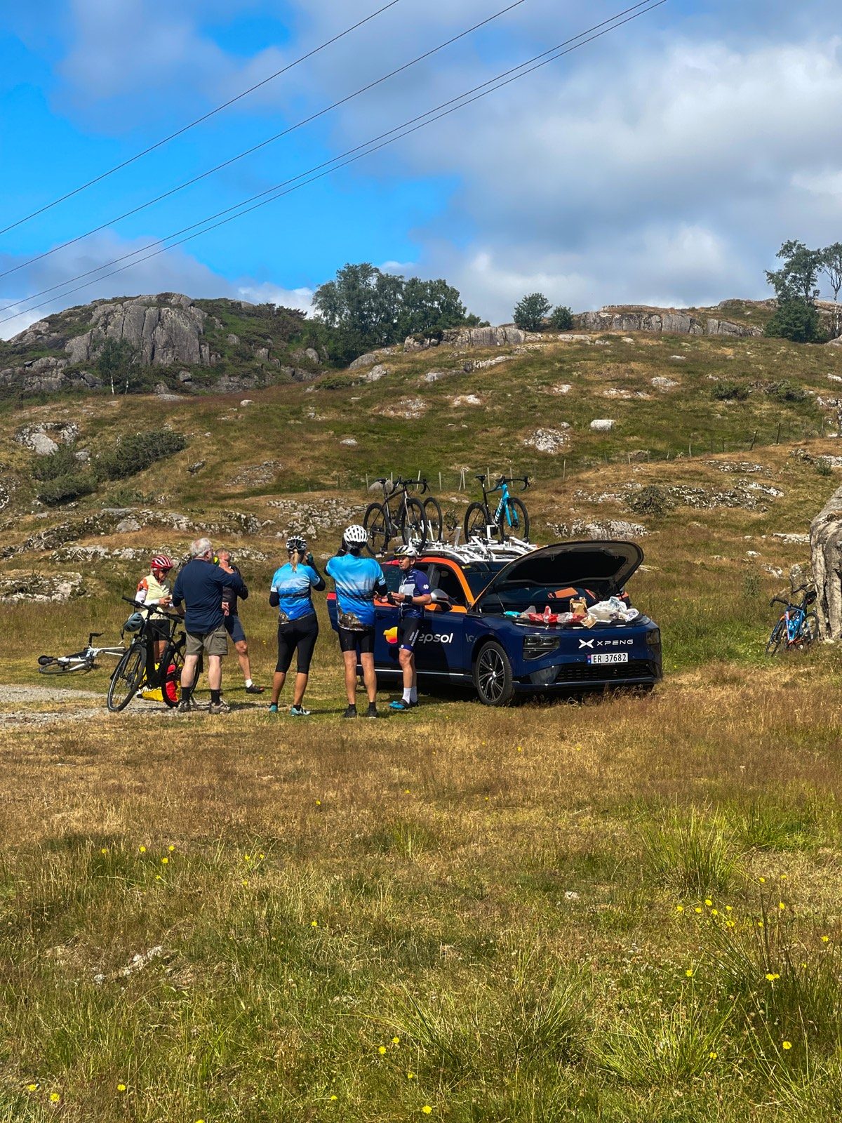 Syklister tar en pause ved en følgebil med sykler og utstyr i fjordlandskapet.
